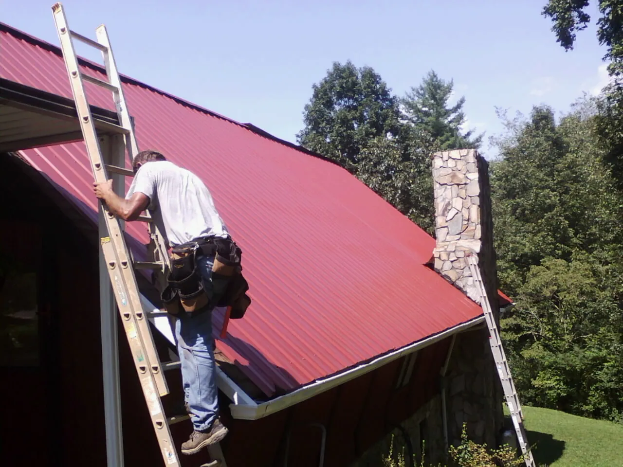 Roofer on a ladder beside a red metal roof and stone chimney — Soffit & Fascia Repair by Roofing Brothers in Wausau